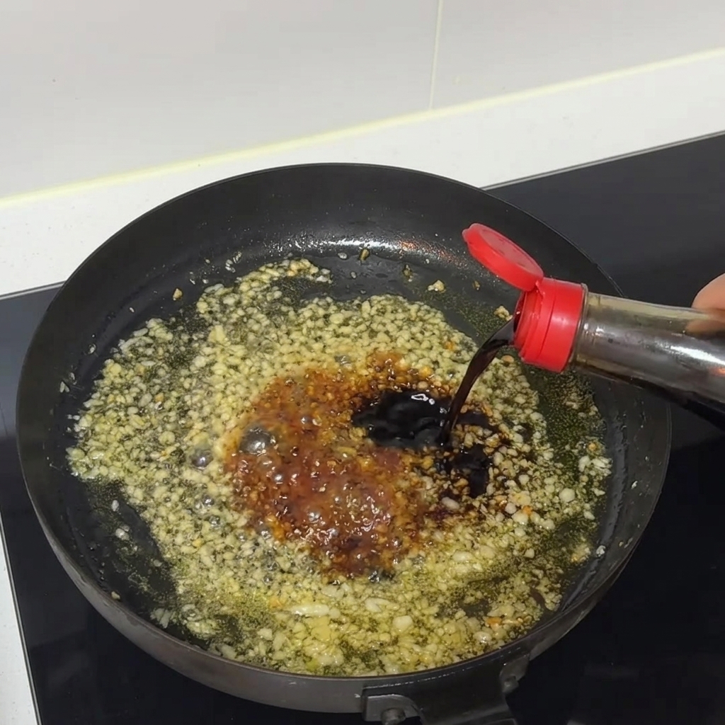 Dunkle Sojasauce fließt aus einer Flasche in eine zischende Pfanne mit gehacktem Knoblauch und Sauce.