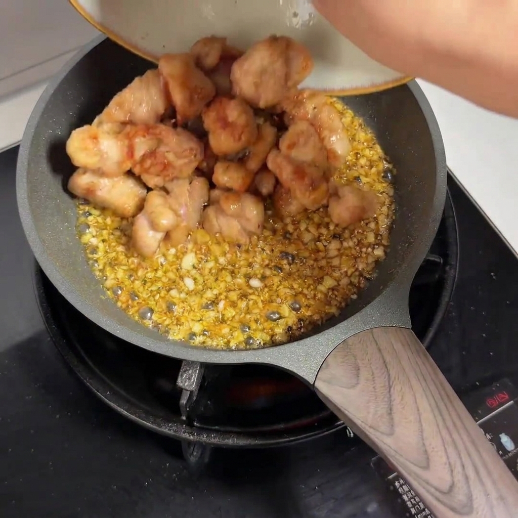 Vorgebratene Hähnchenstücke werden von einem Teller in eine Bratpfanne mit Knoblauchsauce gegeben.