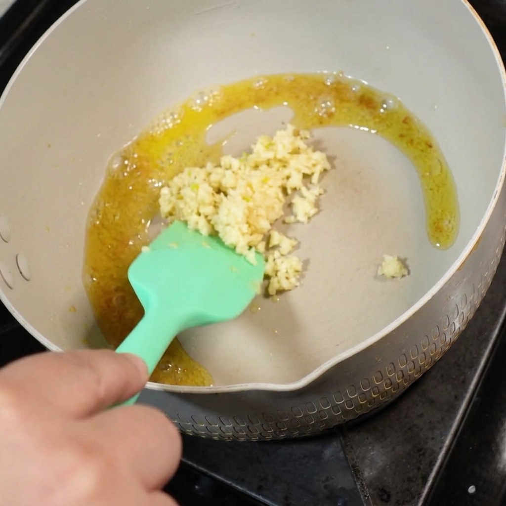 Frischer, gehackter Knoblauch wird zu geschmolzener Butter in eine Pfanne gegeben und mit einem grünen Silikonspatel verrührt.