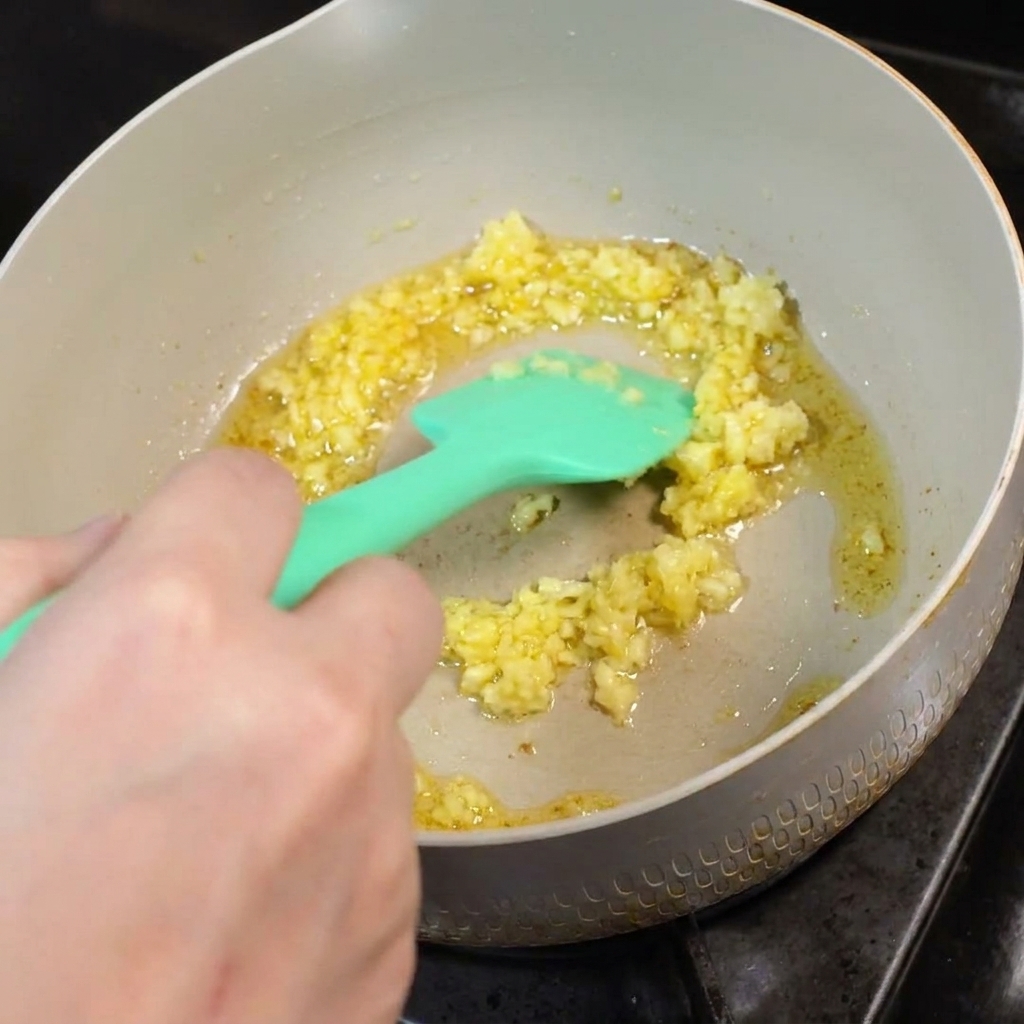Gehackter Knoblauch brät in geschmolzener Butter in einer Pfanne und wird mit einem grünen Silikonspatel umgerührt.