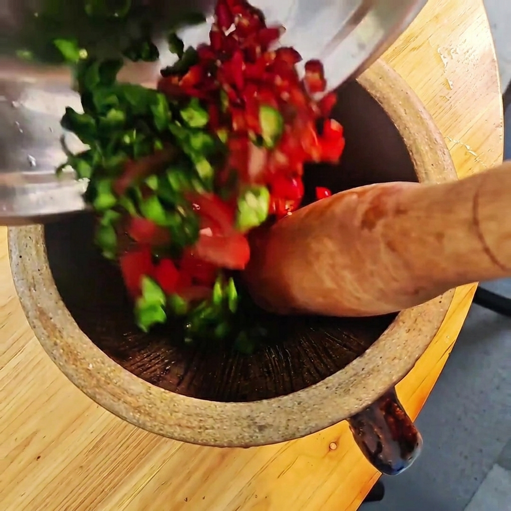Gehackte grüne und rote Chilis sowie Tomaten werden in einem Holzmörser mit einem Stößel zerstoßen.cite: 1
