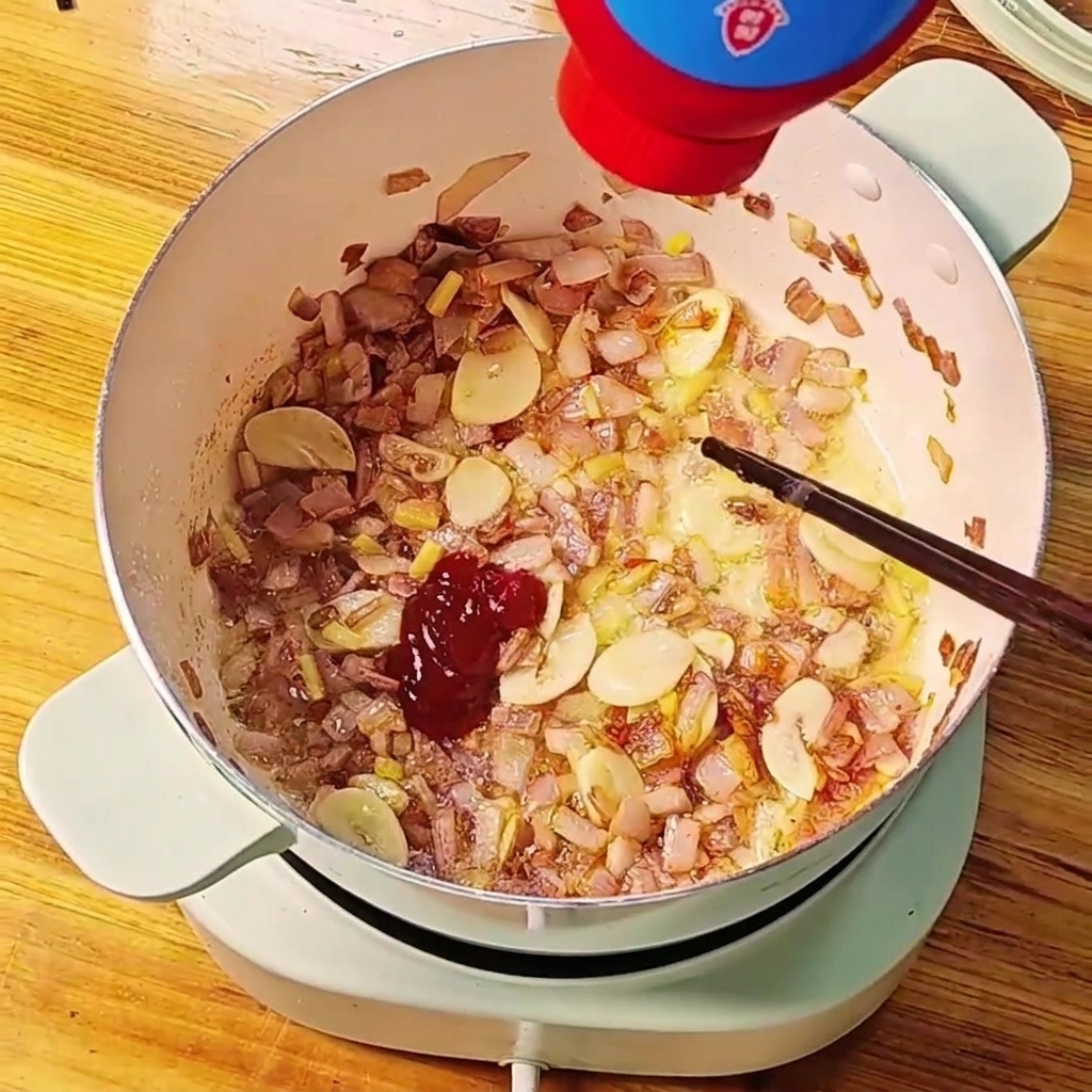 Rotes Tomatenmark wird aus einer Flasche in einen Topf mit angebratenen Zwiebeln und Knoblauch gedrückt.cite: 1