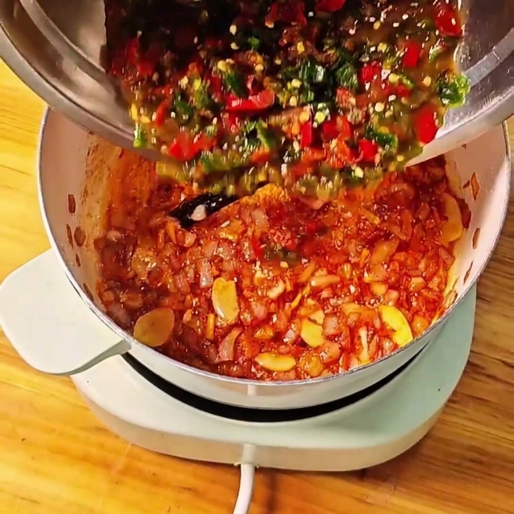 Zerstoßene Tomaten sowie grüne und rote Chilis werden aus einer Metallschüssel in den Kochtopf gegossen.cite: 1