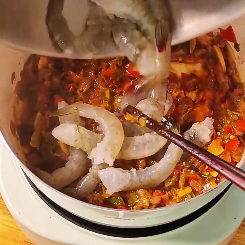 Rohe, graue ganze Garnelen werden in eine köchelnde rote Tomatensauce in einem weißen Topf gegeben.cite: 1