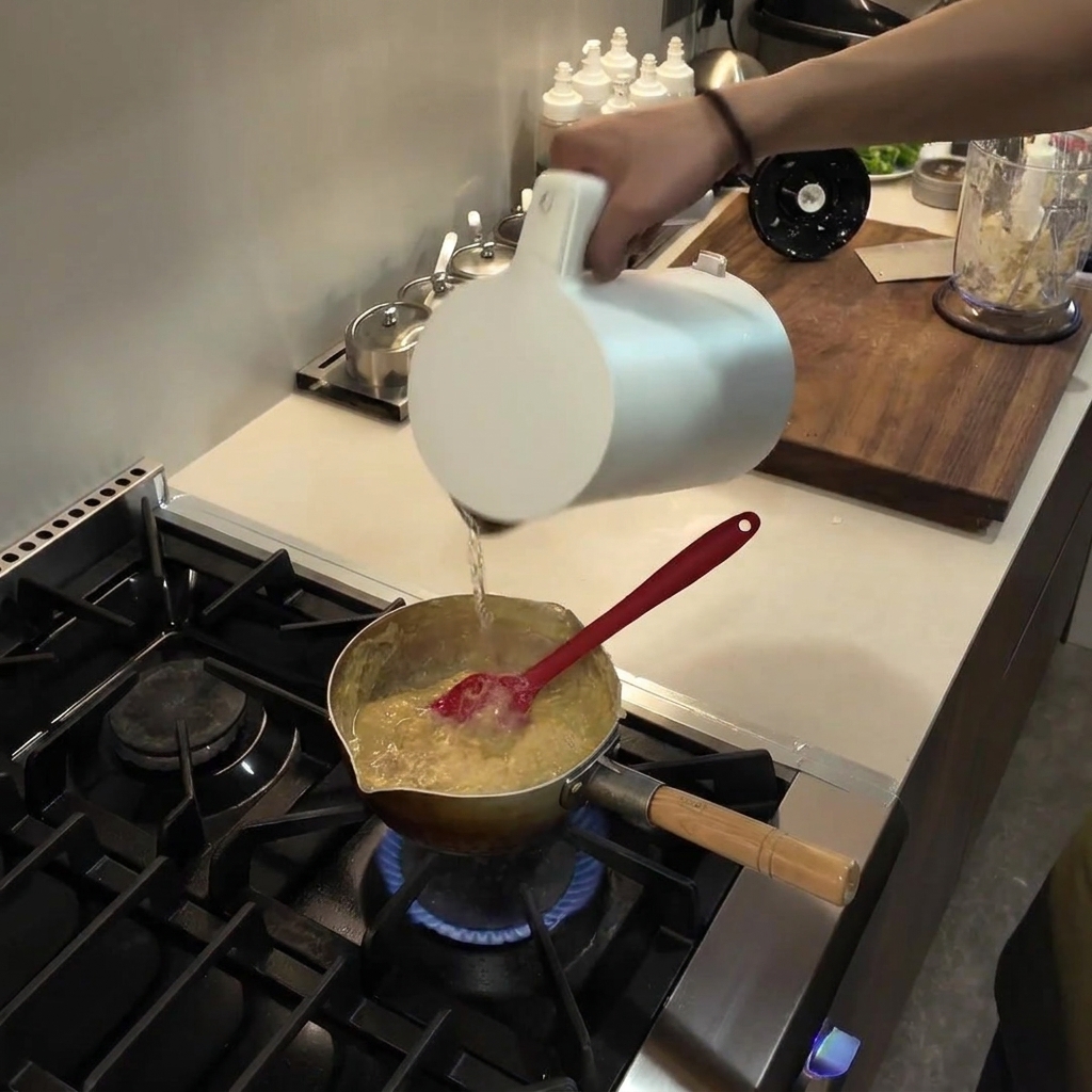 Wasser wird aus einer weißen Karaffe in einen Topf mit einer eindickenden, köchelnden Fufu-Mischung gegossen.