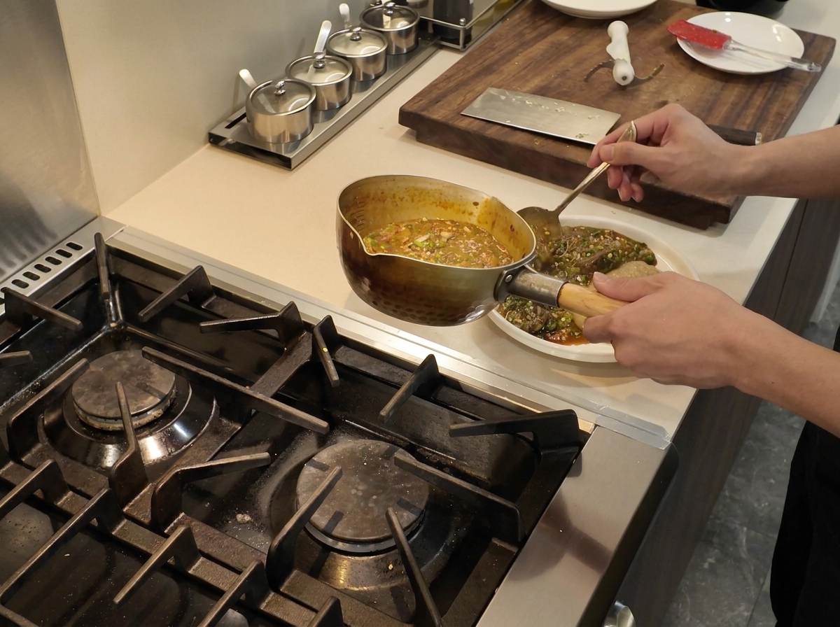 Eine Kelle gießt dampfend heißeren Okra-Fisch-Eintopf neben ein quadratisches Fufu-Stück auf einen weißen Teller.