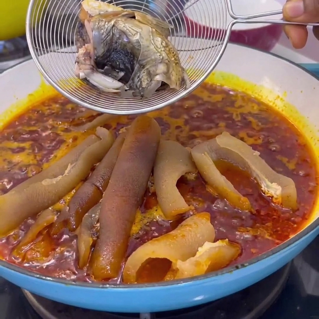 Ein Drahtsieb mit gekochten Fischstücke wird in einen Topf mit köchelnder roter Sauce und Schweinehaut abgesenkt.