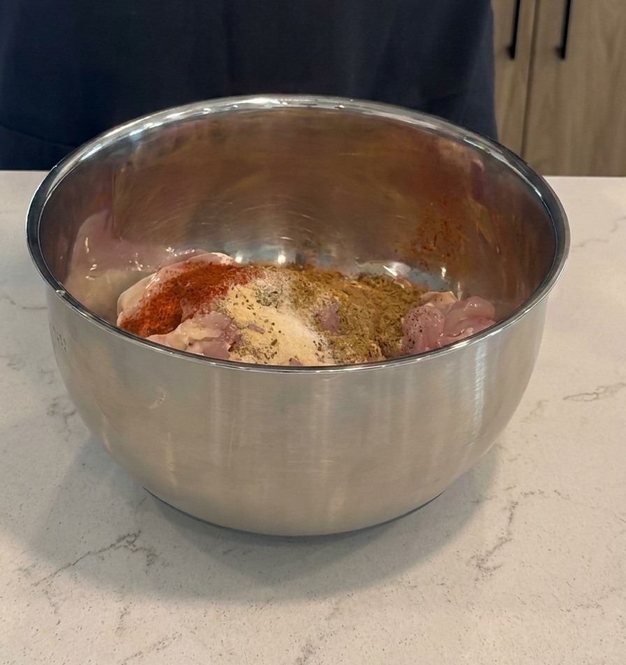 Dunkelrote Marinade und trockene Gewürze werden in einer Edelstahlschüssel über rohe Hähnchenkeulen gegeben.