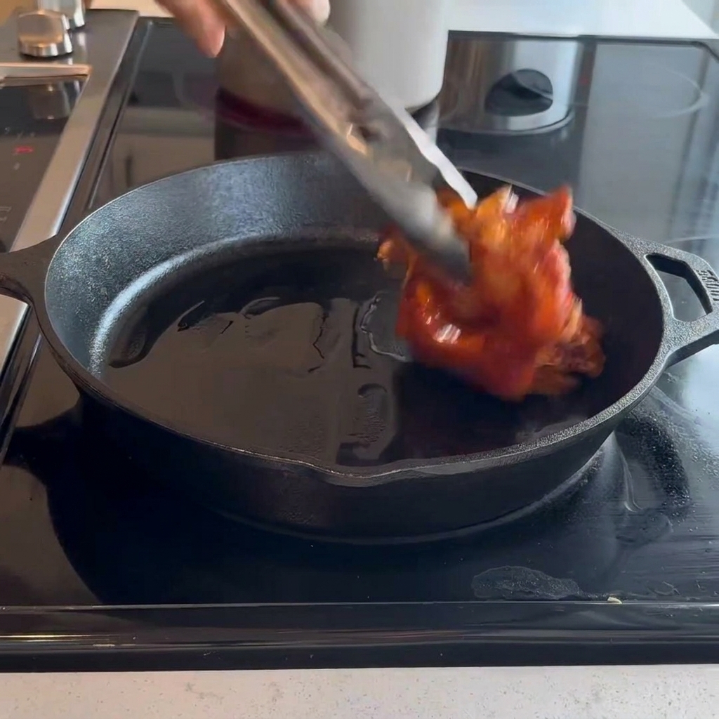 Rohe, marinierte Hähnchenkeulen werden mit einer Metallzange mit der Hautseite nach unten in eine heiße gusseiserne Pfanne gelegt.