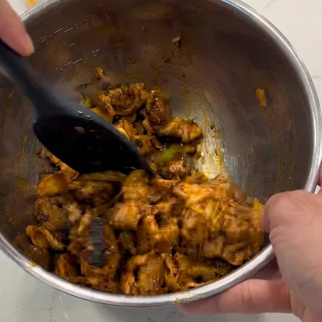 Ein schwarzer Spatel mischt und wendet die Hähnchenwürfel in einer Metallschüssel, um sie mit einer glänzenden, braunen Sauce zu überziehen.