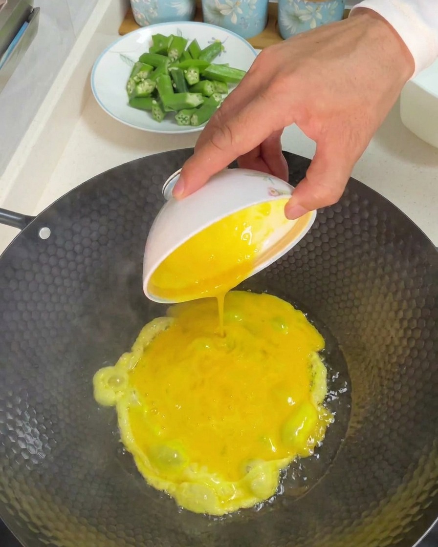 Eine Schüssel mit verquirlten gelben Eiern wird in einen heißen Wok mit Öl gegossen.