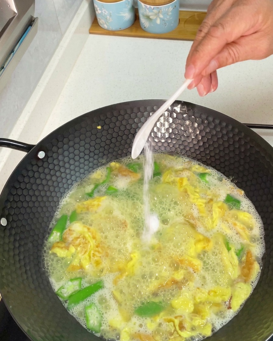 Eine Hand gibt einen Löffel weißes Salz in einen köchelnden Wok mit Okra-Eier-Suppe.