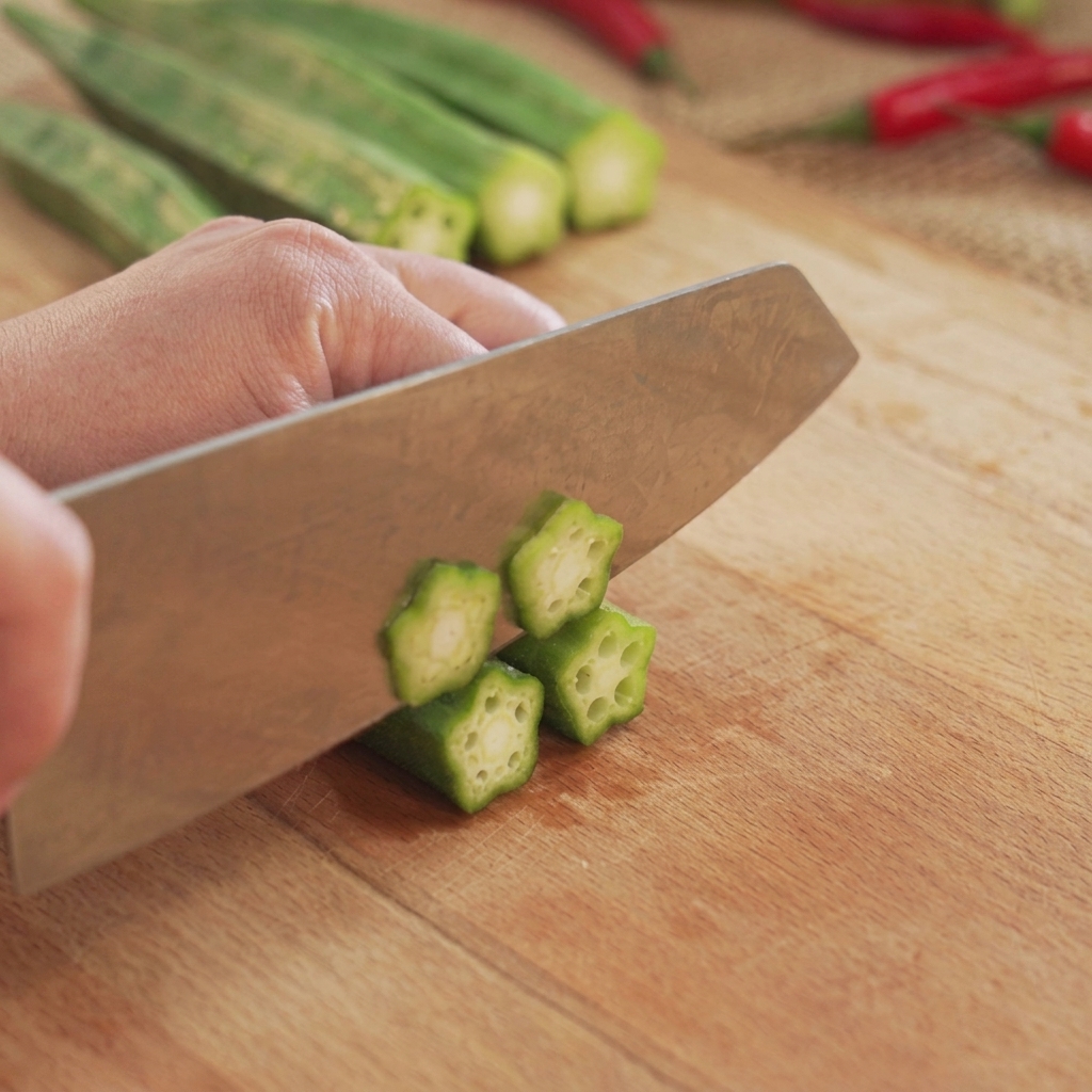 Grüner Okra wird auf einem Holzschneidebrett mit einem Küchenmesser in runde, sternförmige Scheiben geschnitten.