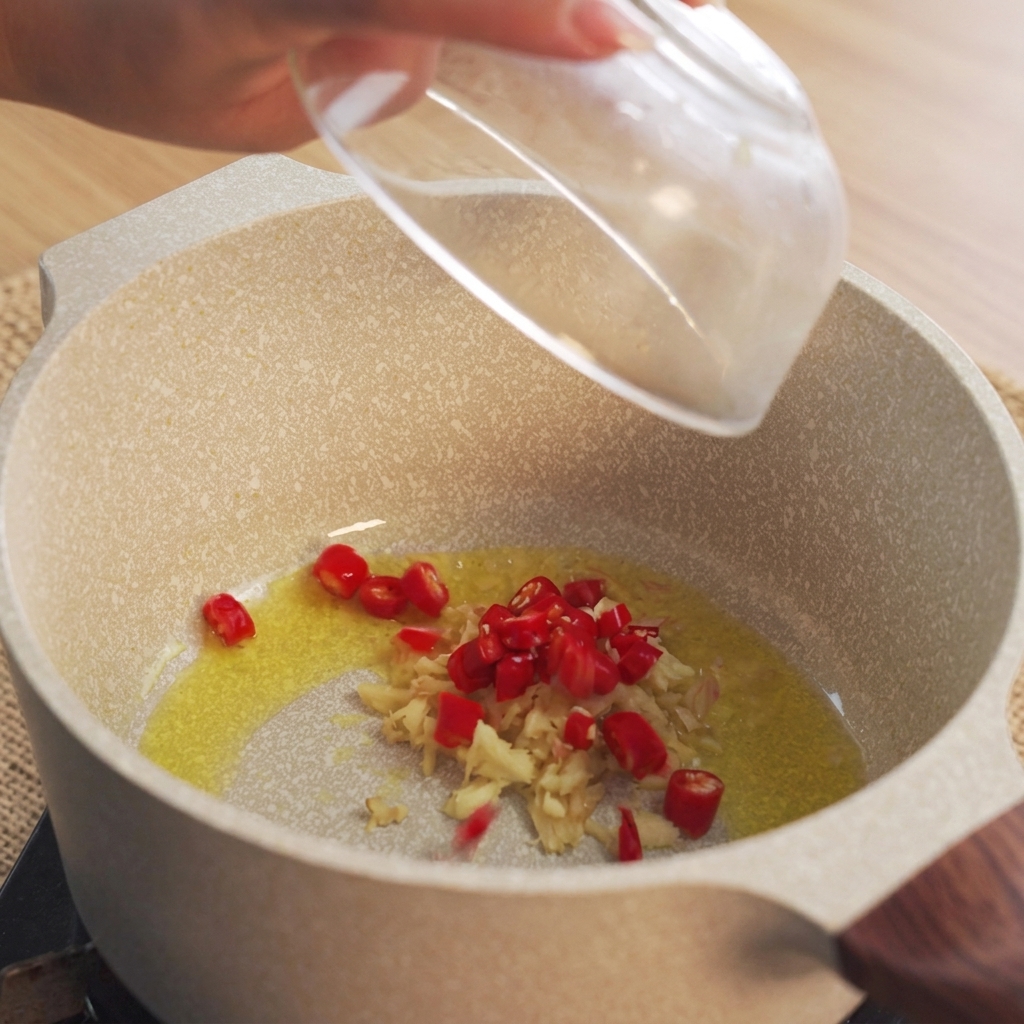Eine Hand gibt gehackte rote Chilischoten in einen Topf mit heißem Öl und Ingwer.