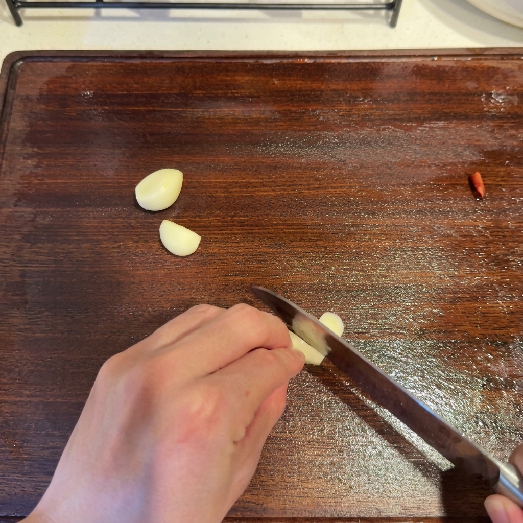 Knoblauchzehen werden mit einem Messer auf einem hölzernen Schneidebrett neben einem Stück roter Chili in Scheiben geschnitten.