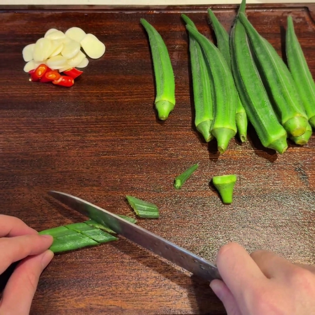 Blanchierte grüne Okra wird mit einem Messer auf einem Holzschneidebrett diagonal in Scheiben geschnitten.
