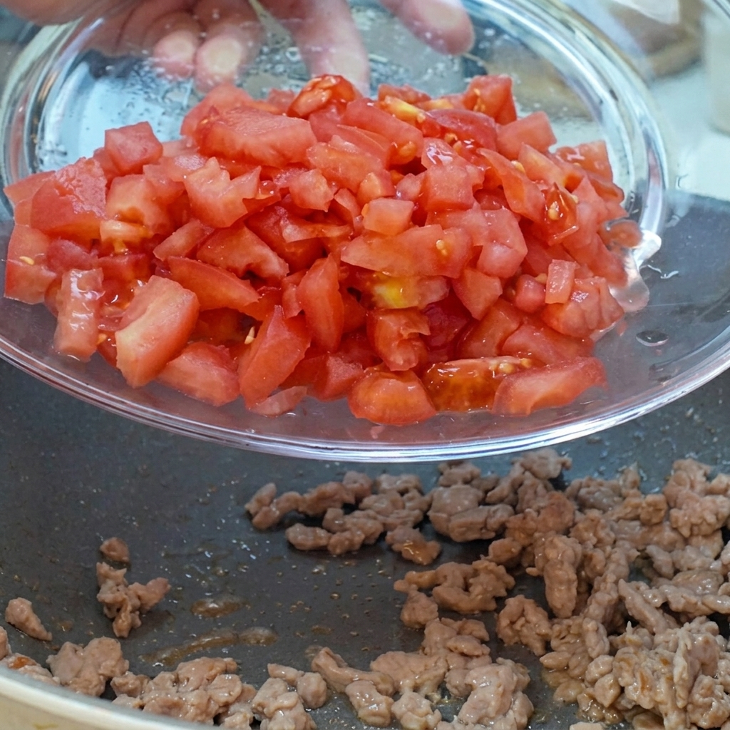 Gewürfelte Tomaten und rote Zwiebeln, die aus einer Glasschüssel in die Pfanne zum Fleisch gegeben werden.