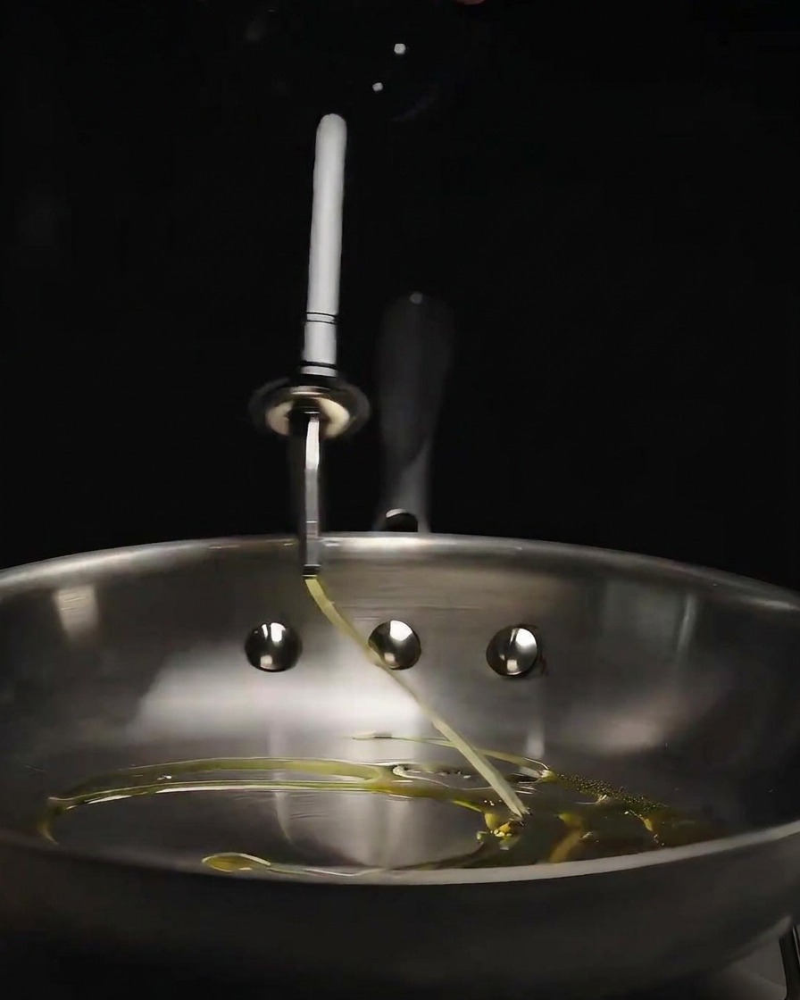 Olivenöl wird aus einer Glasflasche in eine glänzende Bratpfanne aus Edelstahl gegossen.