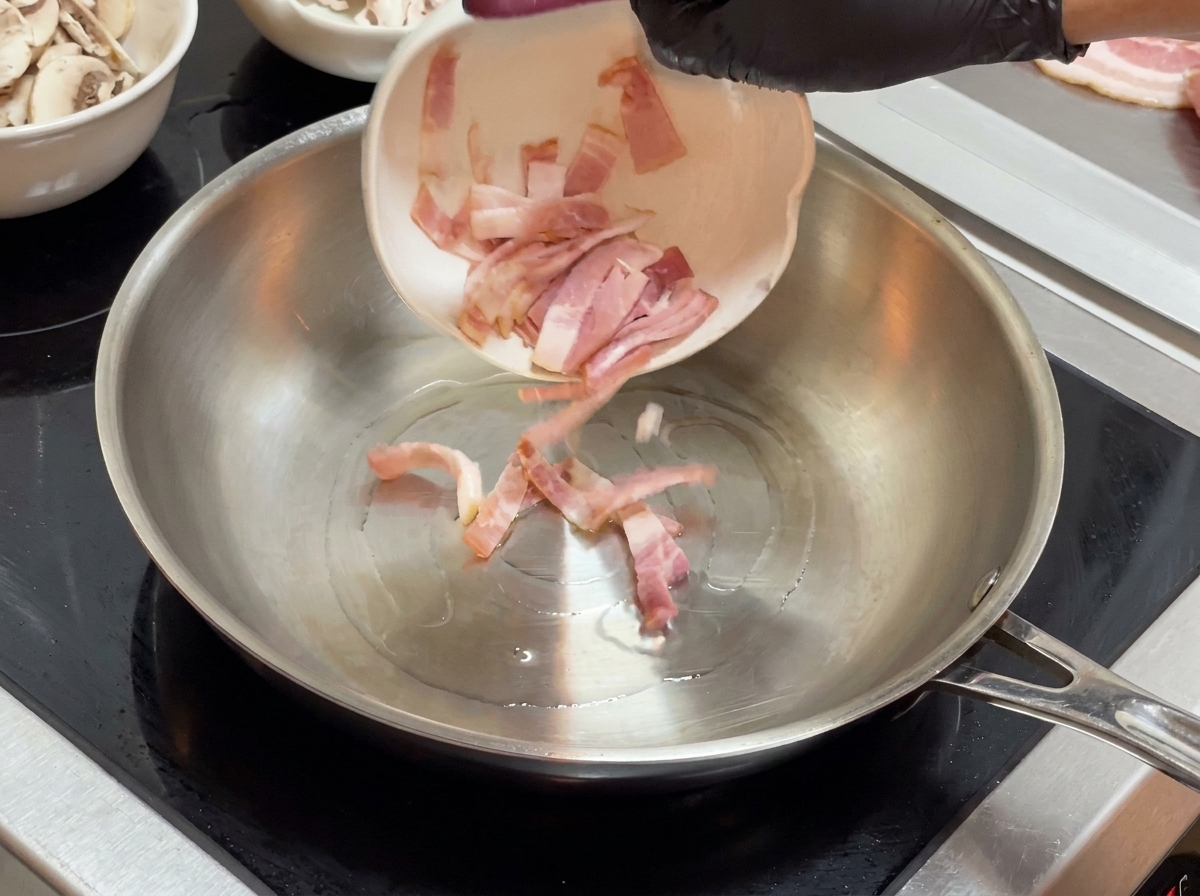 Speckstücke werden aus einer kleinen weißen Schüssel in eine Bratpfanne aus Edelstahl gegeben.