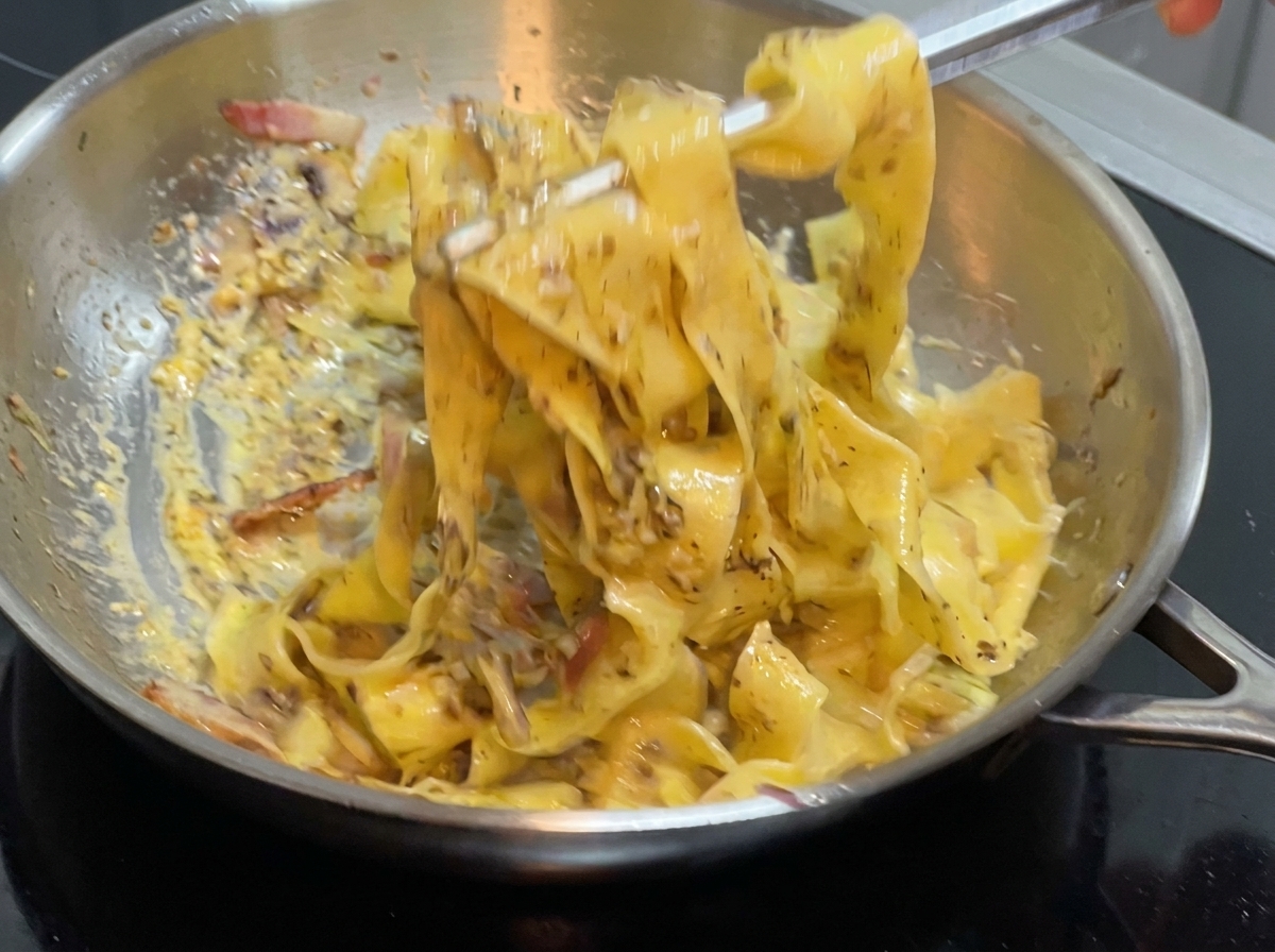 Eine Zange mischt breite Pasta und Sauce in einer Edelstahlpfanne auf dem Herd.