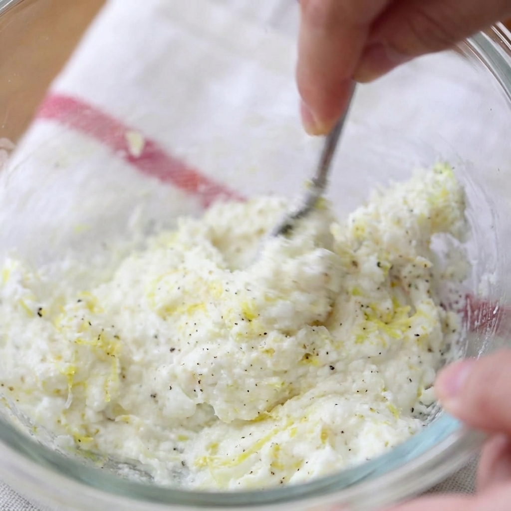 Eine Hand vermengt Frischkäse, Zitronenabrieb und schwarzen Pfeffer in einer Glasschüssel mit einer Gabel.