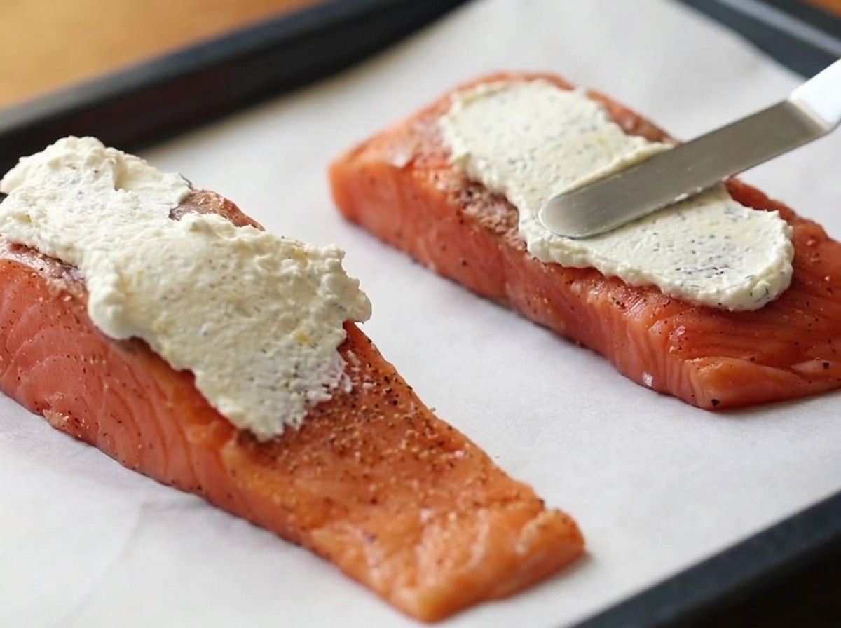 Ein Metallspatel verstreicht eine dicke Schicht der weißen Frischkäsemischung auf zwei rohen Lachsfilets, die auf einem mit Backpapier ausgelegten Backblech liegen.
