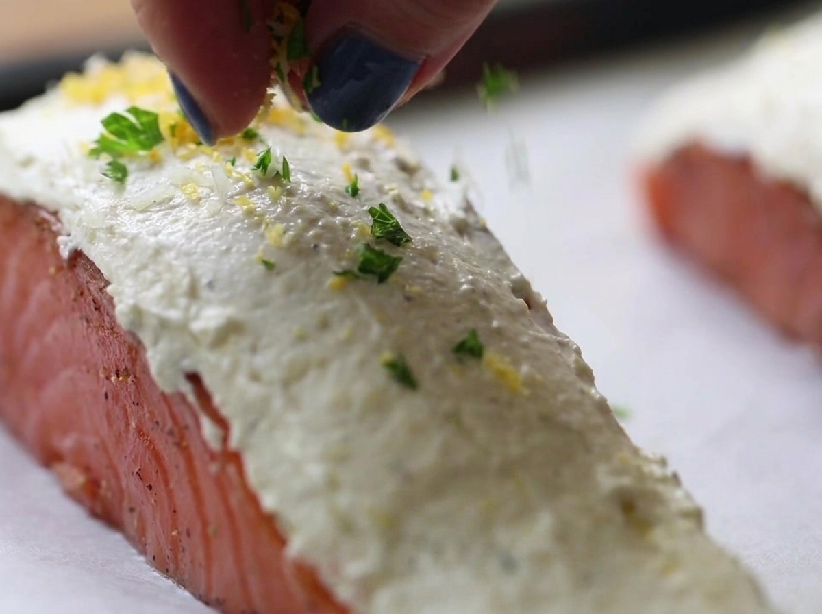Finger streuen eine grün-gelbe Kräuter-Paniermehl-Mischung über rohe Lachsfilets, die mit weißem Frischkäse bestrichen sind.