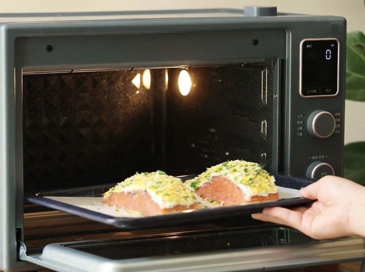 Eine Hand schiebt ein dunkles Backblech mit zwei panierten Lachsfilets in einen kleinen, hell erleuchteten Tischbackofen.