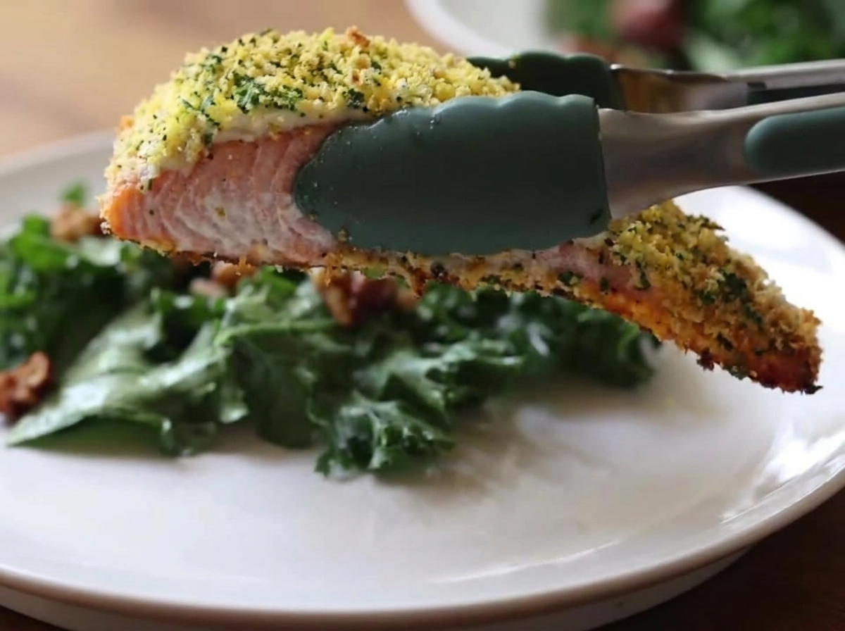 Eine grüne Küchenzange aus Silikon hält ein Stück gebackenen Lachs mit einer goldbraunen, knusprigen Kruste über einen weißen Teller mit grünem Grünkohlsalat.