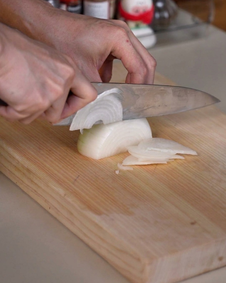 Eine Zwiebel wird auf einem Holzschneidebrett mit einem scharfen Küchenmesser sorgfältig in dünne Streifen geschnitten.