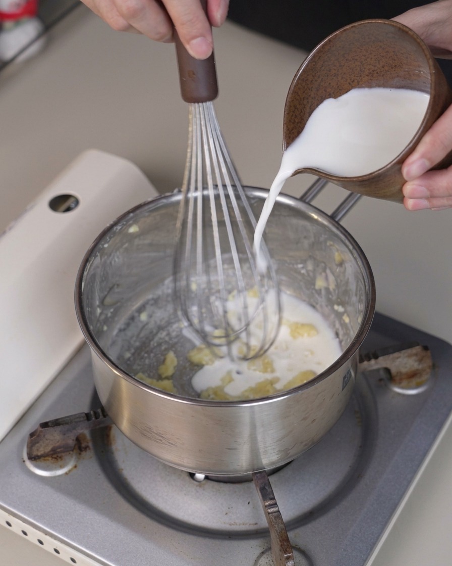 Kalte Milch wird aus einer kleinen Kanne in einen Topf gegossen, während ein Schneebesen die Mischung verrührt.