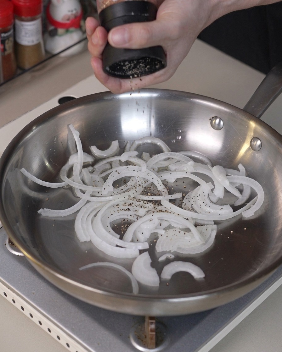 In Scheiben geschnittene Zwiebeln werden mit Pfeffer gewürzt und in einer Edelstahlpfanne geschwenkt.