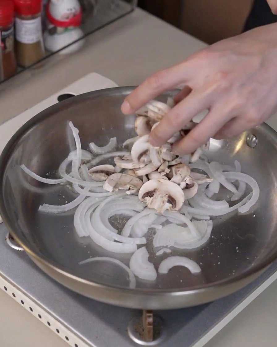 In Scheiben geschnittene Champignons werden in die Pfanne zu den Zwiebeln gegeben, um zu garen und Feuchtigkeit abzugeben.