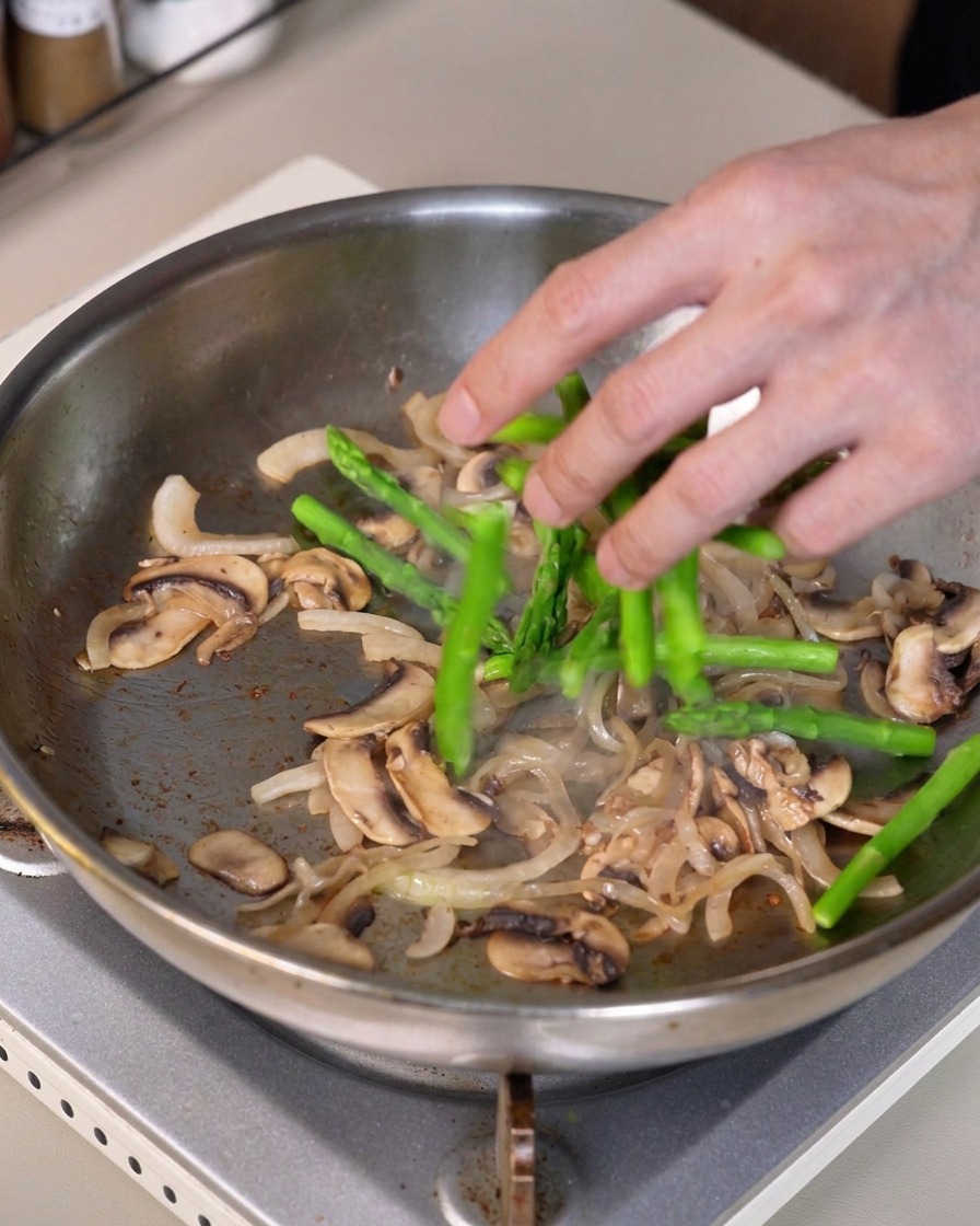 Grüne Spargelstücke werden in die Pfanne mit den angebratenen Zwiebeln und Pilzen gemischt.