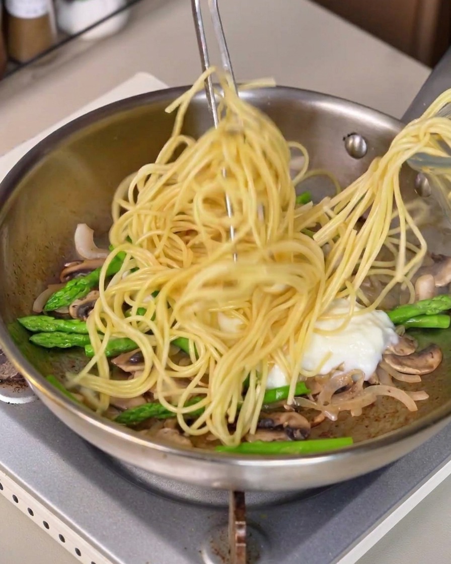 Lange Spaghetti-Pasta wird mit einer Zange angehoben und in die Pfanne mit der weißen Soße und dem Gemüse gemischt.