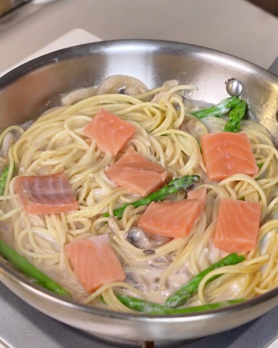 Frische Lachsstücke werden über einer Pfanne mit cremiger Pasta und Spargel verteilt.