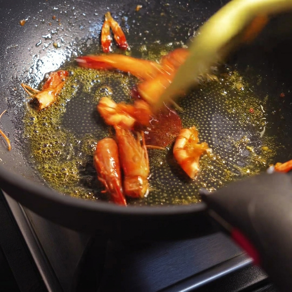 Garnelenköpfe und -schalen werden in einer Pfanne angebraten und geben ein leuchtend orangerotes Öl ab.