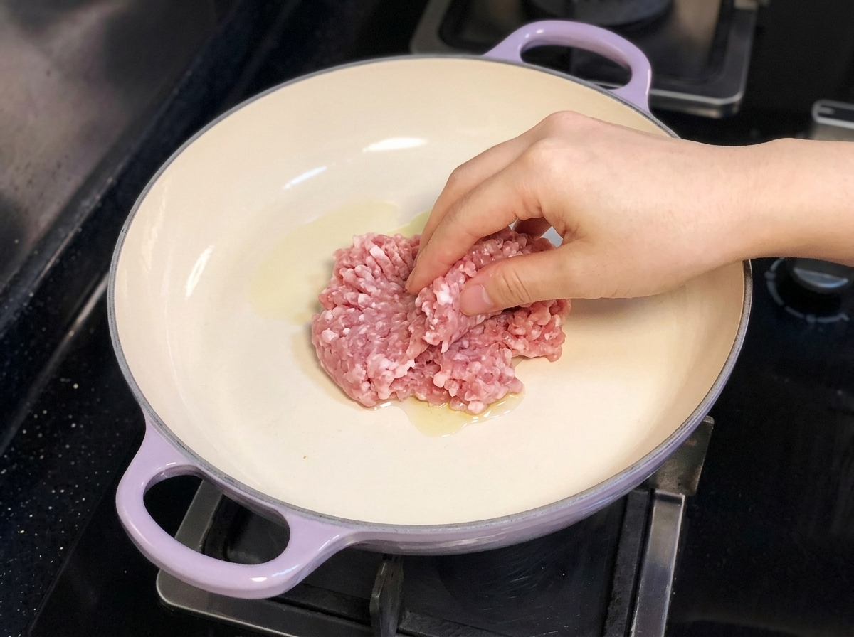 Eine Hand gibt Schweinehackfleisch in eine lila griffige Pfanne mit Olivenöl auf einem Herd.
