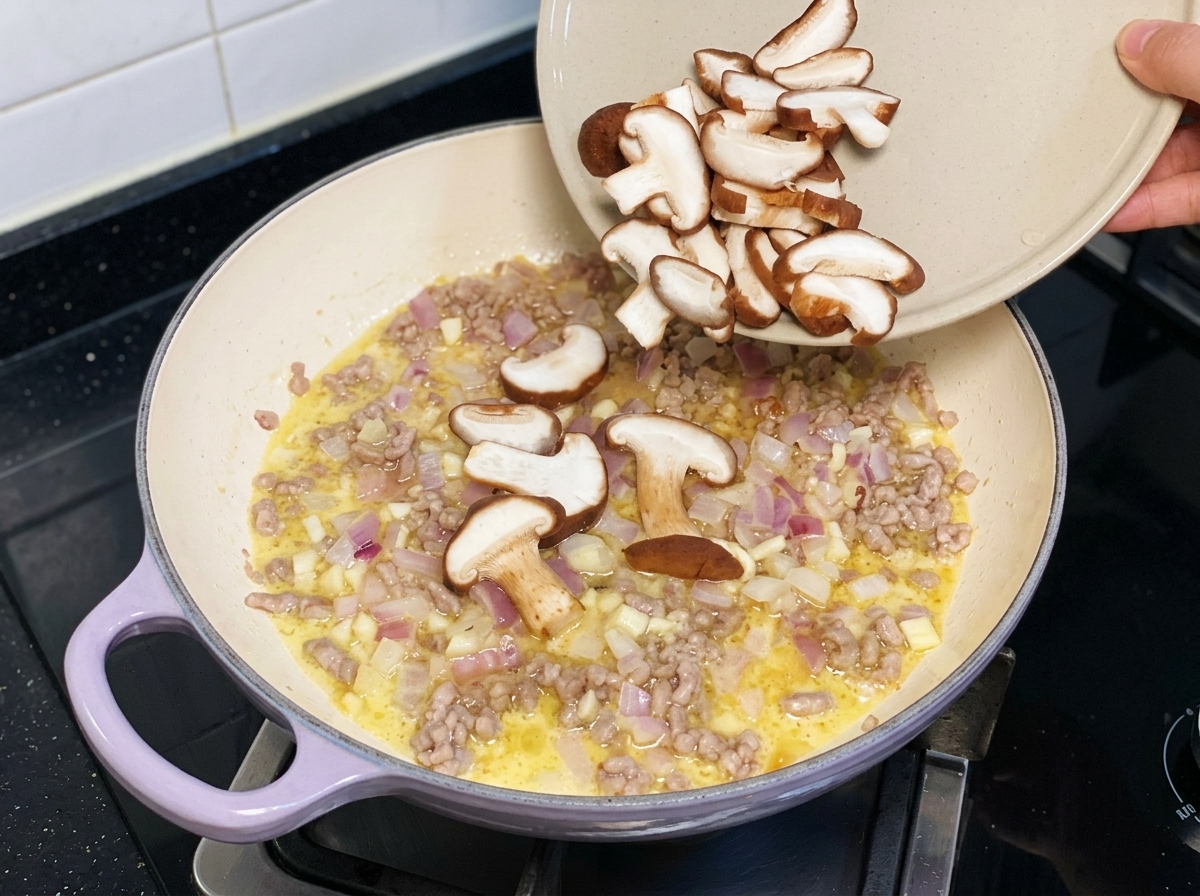 In Scheiben geschnittene frische Shiitake-Pilze werden von einem Teller in eine Pfanne mit Hackfleisch und Zwiebeln gegeben.