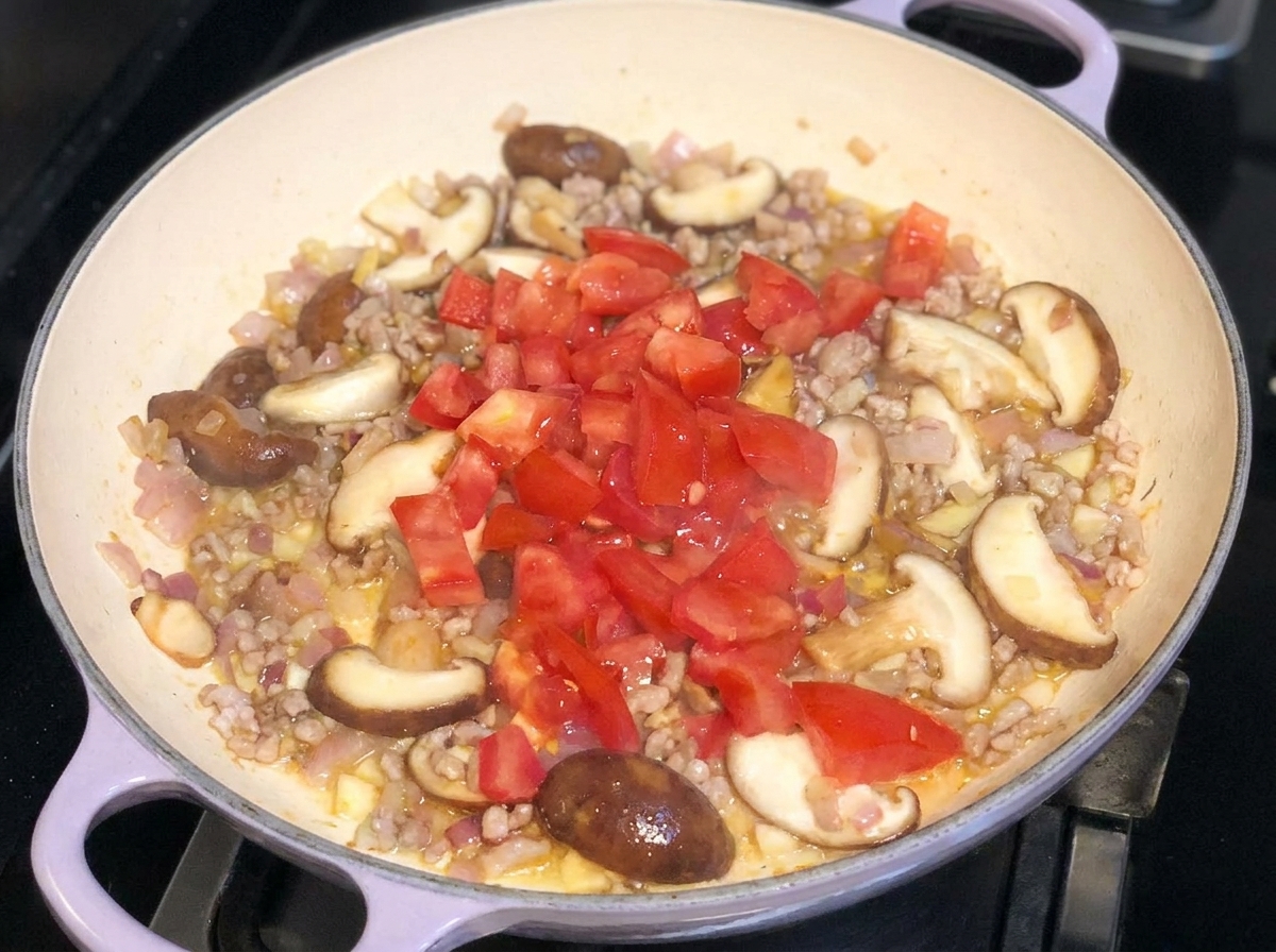 Frisch gewürfelte Tomaten werden auf eine angebratene Mischung aus Fleisch, Zwiebeln und Pilzen in einer Pfanne gegeben.