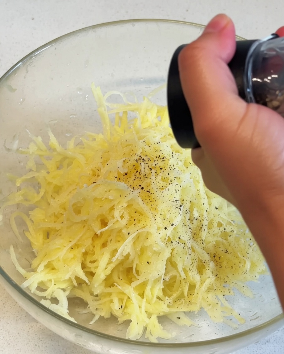 Geraspelte gelbe Kartoffeln in einer Glasschüssel, die mit schwarzem Pfeffer aus einer Handmühle gewürzt werden.