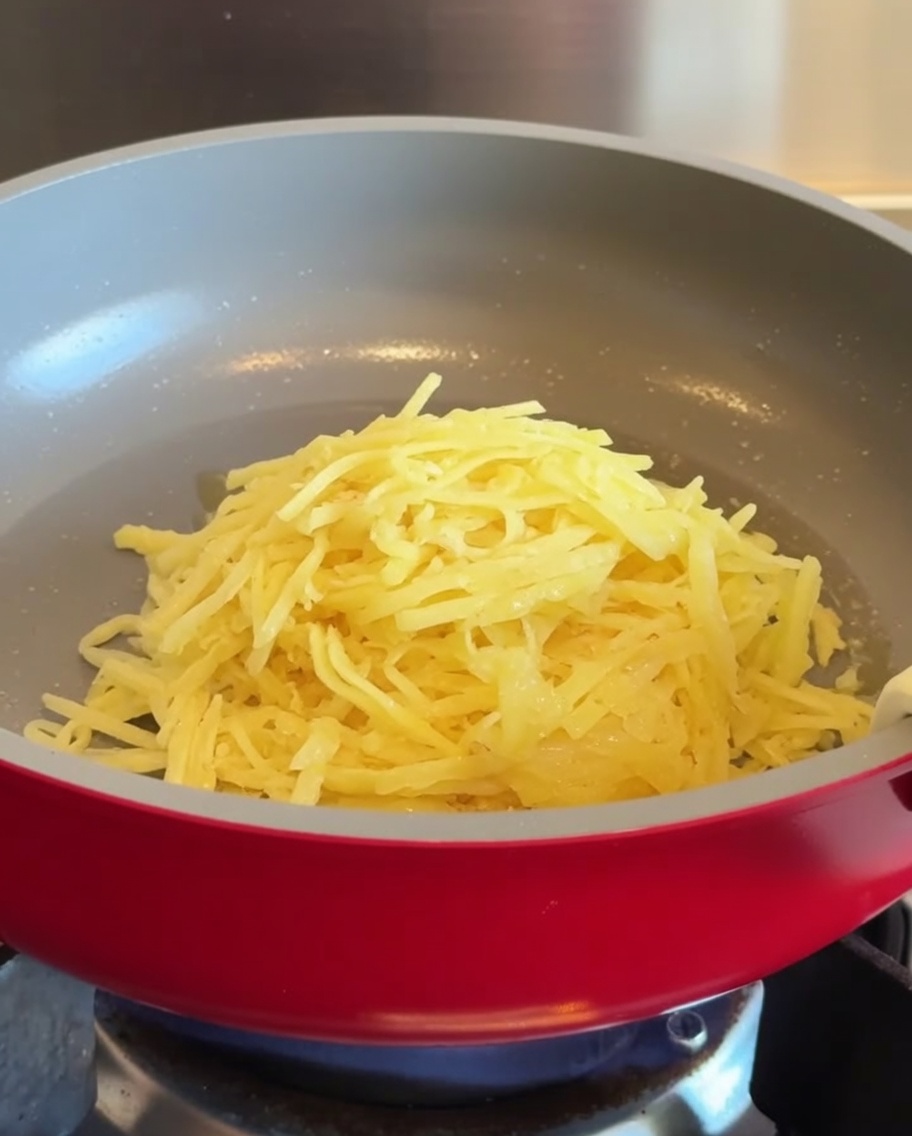 Ein Haufen geraspelter Kartoffeln in der Mitte einer erhitzten roten Antihaft-Pfanne.
