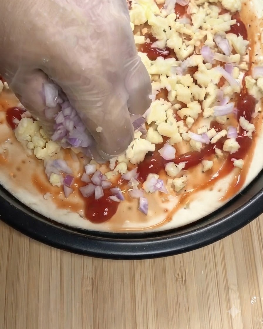 Eine Hand mit Handschuh streut gewürfelte rote Zwiebeln und geriebenen Mozzarella auf einen Pizzaboden mit Tomatensoße.