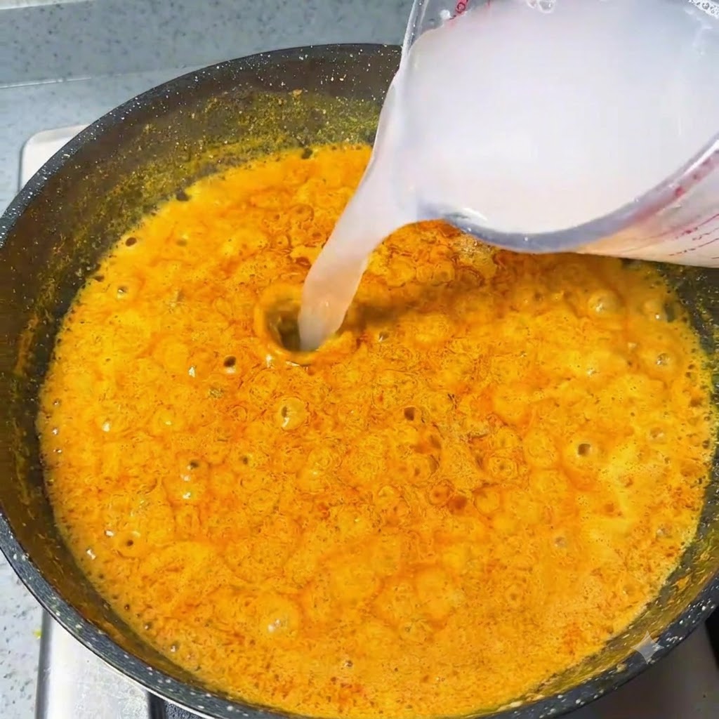 Wasser wird aus einem Messbecher in eine blubbernde orangefarbene Currysoße gegossen.
