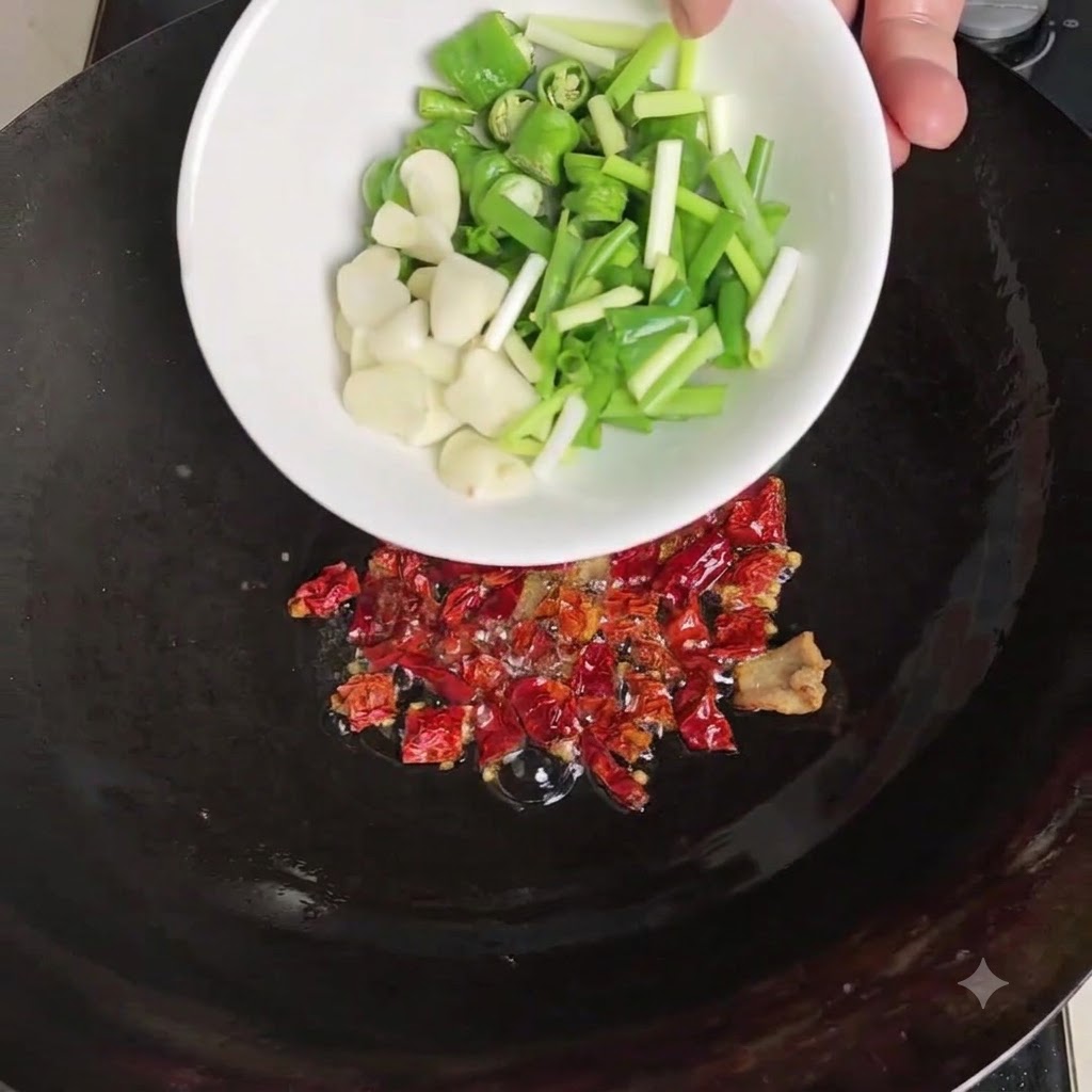 Eine weisse Schüssel giesst geschnittene grüne Chilis, geschälten Knoblauch und Frühlingszwiebeln über die angerösteten getrockneten Chilis im Wok.