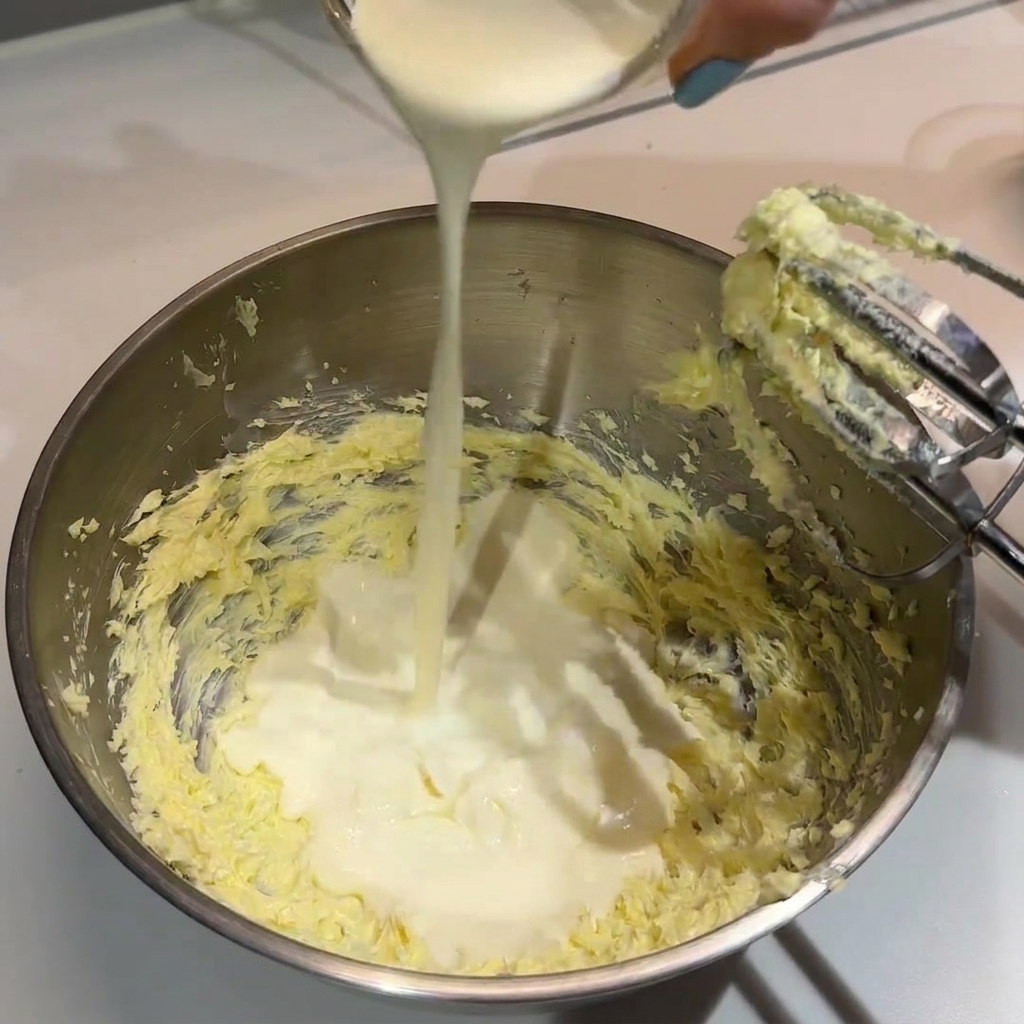 Schlagsahne wird aus einem kleinen Glasbehälter in die Rührschüssel mit geschlagener Butter gegossen.