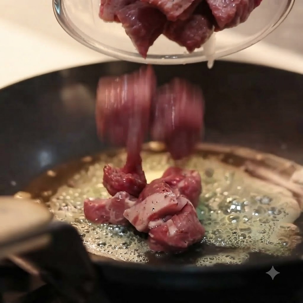 Stücke von gewürztem rohem Rindfleisch werden aus einer Glasschüssel in eine heiße Pfanne mit sprudelnder, geschmolzener Butter gefüllt.