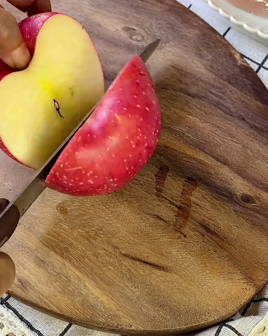 Ein Apfel wird auf einem hölzernen Schneidebrett mit einem silbernen Messer in kleine Stücke geschnitten.