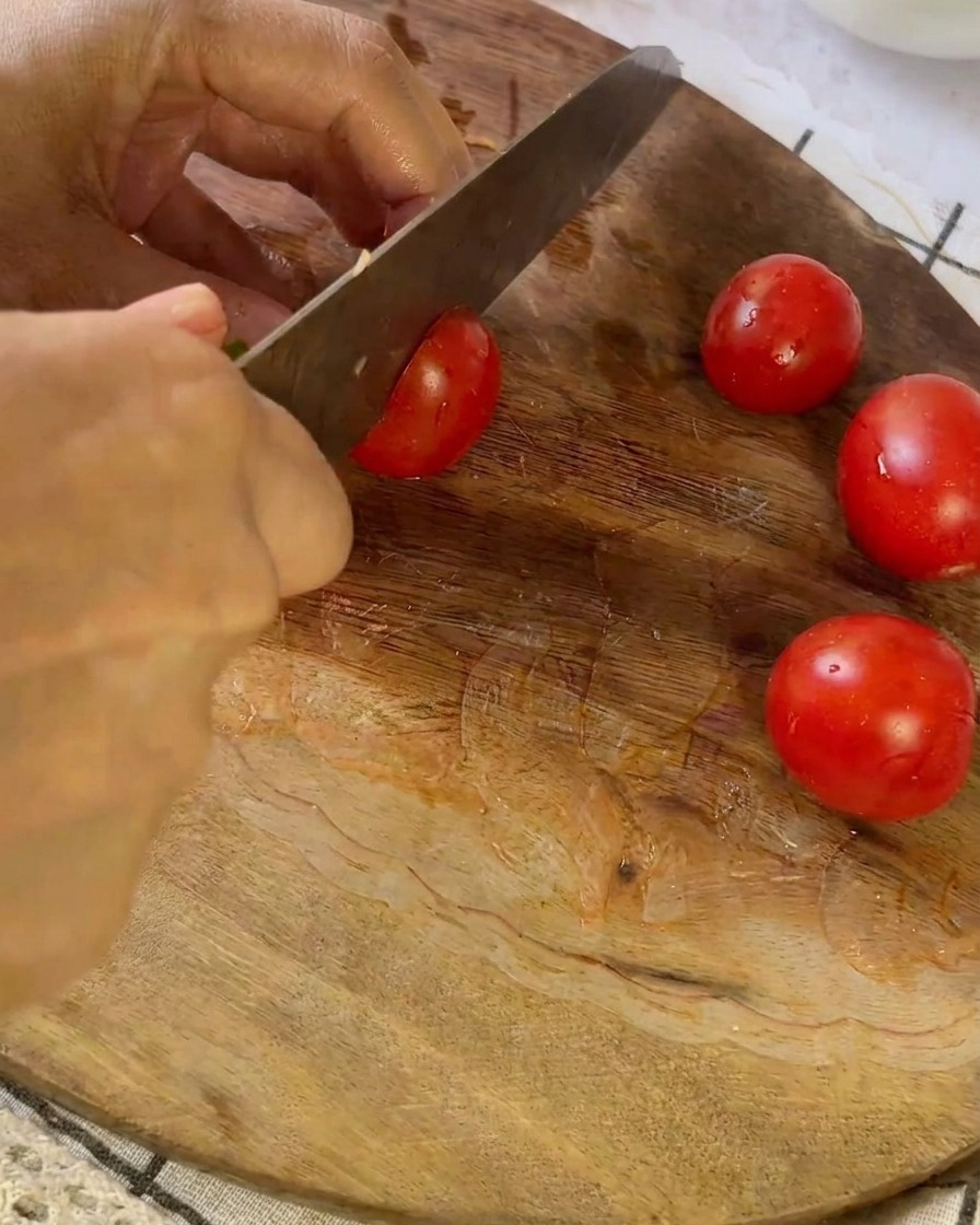 Ein Messer schneidet eine kleine rote Tomate auf einem Holzschneidebrett neben anderen halbierten Tomaten in der Hälfte durch.