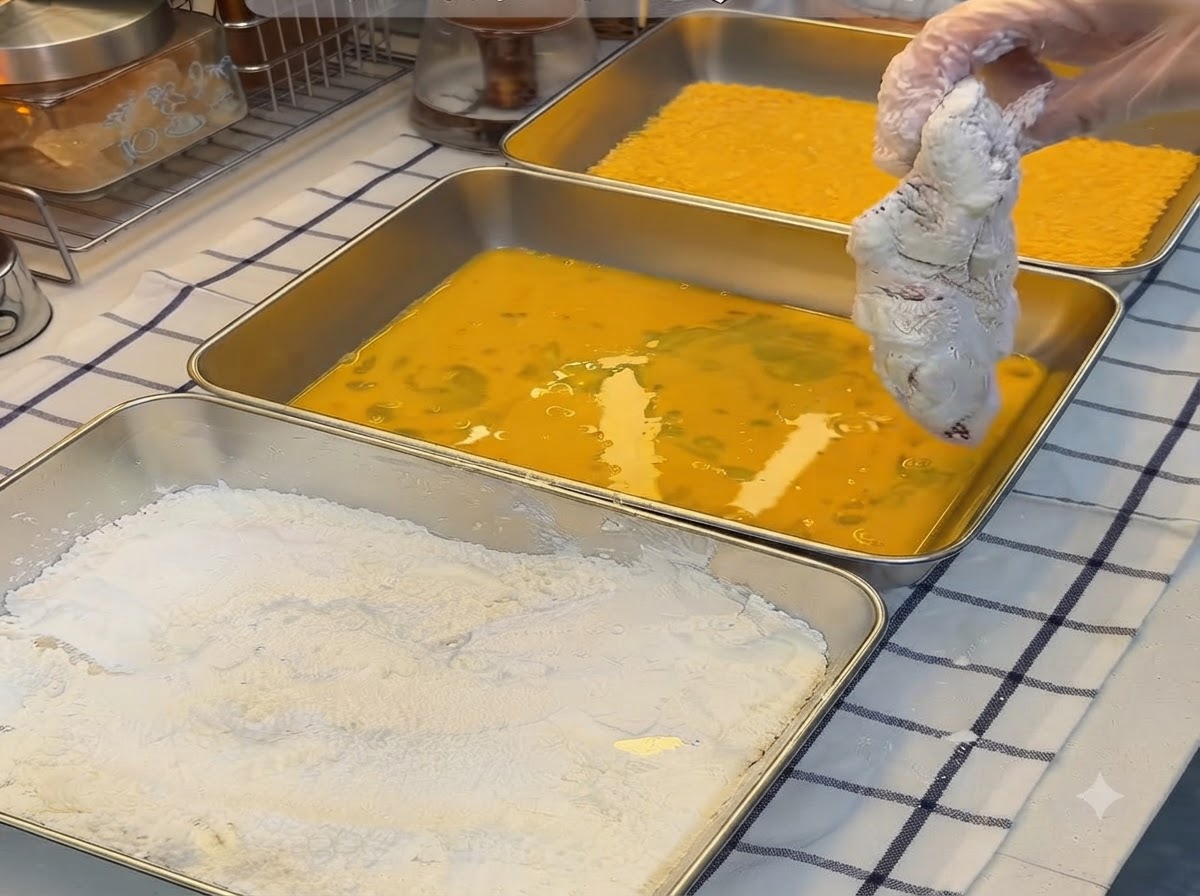 Ein mit Stärke paniertes Schweinekotelett wird in eine Schale mit verquirlten gelben Eiern getaucht.